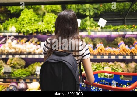 Rückansicht einer nicht erkennbaren Kundin mit Rucksack und Warenkorb, die frisches Gemüse in den Regalen im Supermarkt wählt Stockfoto