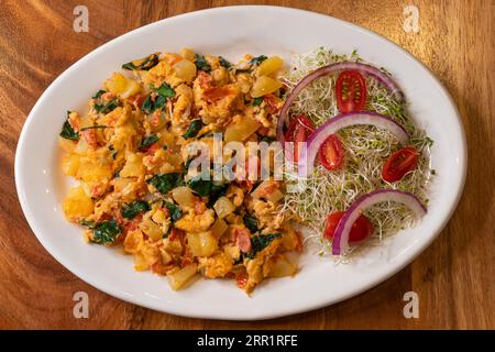 Blick von oben auf appetitliche Spiegeleier mit grüner Blattsauce und Tomaten, Zwiebeln garniert auf Sprossen, serviert auf weißem Marmorteller auf hölzerner Oberfläche Stockfoto