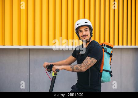Seitenansicht des lächelnden Lieferers im Helm, der auf einem elektrischen Tretroller auf einer Asphaltstraße in der Nähe der Hausmauer in der Stadt steht Stockfoto