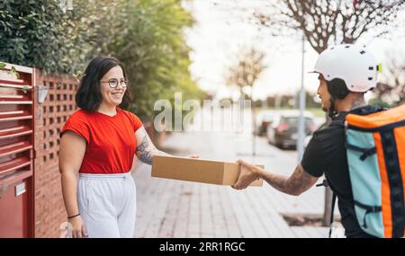 Fröhliche junge Frau in lässiger Kleidung, die Paket vom Liefermann im Schutzhelm auf dem Bürgersteig erhält Stockfoto