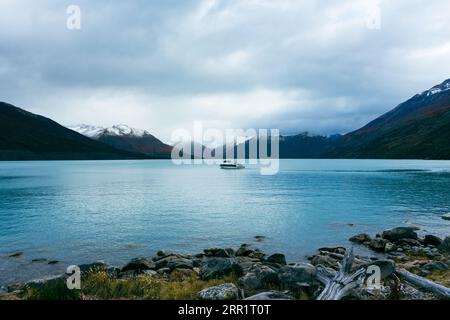 Malerischer Blick auf das Boot auf dem türkisfarbenen See in der Nähe von felsigen Gletscherbergen und steinigen Ufern mit Holzholz gegen bewölkten Himmel im Winter in Perito Moreno Arge Stockfoto