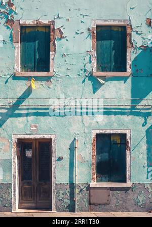 Das Äußere eines alten, leeren, verlassenen Gebäudes mit verschlossenen Fenstertüren und einer schäbigen, von Zement abblätternden blauen Wand mit Pipeline im Sonnenlicht in Venedig Stockfoto