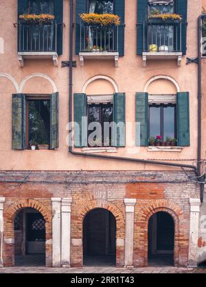 Malerische Fassade eines alten, farbenfrohen Gebäudes mit Blumen auf Fenstern und Balkonen über bogenförmigen Eingängen auf der Straße von Venedig Stockfoto