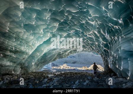 Rückansicht eines anonymen Touristen, der in der Nähe des Fahrrads unter dem Loch steht, während er die Eishöhle in der Schweiz am Wintertag erkundet Stockfoto