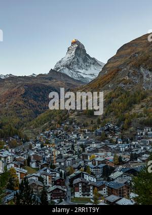 Von oben atemberaubende Aussicht auf ein kleines Dorf inmitten grüner Hügel und schneebedeckter Gipfel unter einem blauen Nachmittagshimmel in der Schweiz Stockfoto