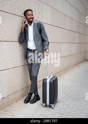 Lächelnder afroamerikanischer männlicher Unternehmer im formellen Anzug im Rucksack, der schaut, während er auf dem Handy in der Nähe des Gebäudes spricht Stockfoto
