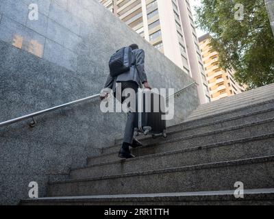 Von unten, Rückansicht des unerkennbaren afroamerikanischen Mannes in formeller Kleidung, der mit Koffer und Rucksack die Treppe hinaufgeht Stockfoto