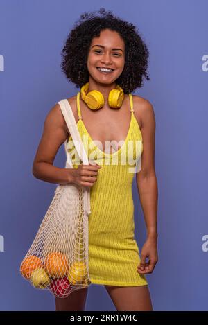 Positive junge hispanische Frau in trendigem gelben Outfit lächelnd und schauend in die Kamera, während sie wiederverwendbare Netztasche voller frischer Früchte gegen Blau trägt Stockfoto