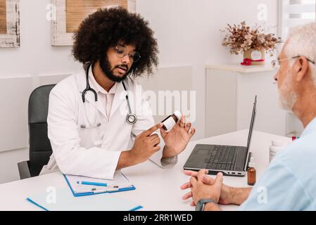 Seriöser bärtiger männlicher Arzt mit Stethoskop und Brille, der am Tisch sitzt und dem Patienten im Krankenhaus eine Flasche Pillen zeigt Stockfoto