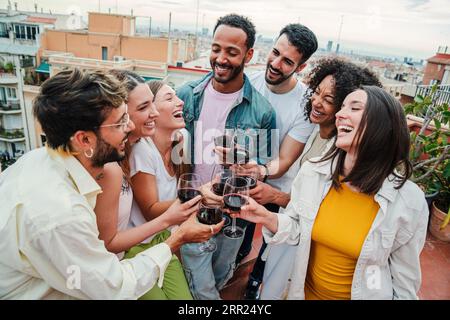 Gruppe junger, fröhlicher Freunde, die lächeln und Rotweingläser auf der Geburtstagsfeier auf dem Dach toasten. Glückliche Erwachsene, die Spaß haben, wenn sie am Wochenende eine Weinbrille klatschen. Freunde, die Schnapsgetränke halten. Hochwertige Fotos Stockfoto