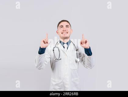 Gutaussehender Arzt zeigt eine Werbung auf. Glücklicher Arzt, der isoliert nach oben schaut und zeigt. Lächelnder Arzt zeigt eine Promo auf Stockfoto