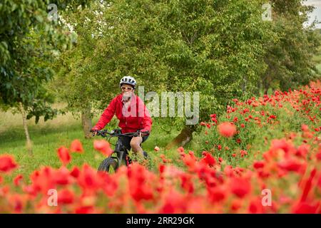 Nette, aktive Seniorin, die ihr Elektrofahrrad in einem riesigen Feld blühender roter Mohnblumen fuhr Stockfoto