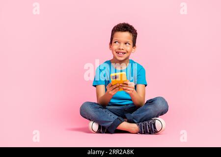 Foto in voller Länge von einem aufregenden, träumenden kleinen Jungen, der ein blaues T-Shirt trägt und ein modernes Gadget textet, das leer aussieht und einen isolierten rosa Farbhintergrund aufweist Stockfoto