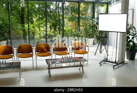 Konferenzraum mit Sesseln, Stühlen und Leinwand. Ein Treffpunkt in einem Glashaus mit Glaswand. Garten für eine Veranstaltung. Innenraum eines leeren Raums für Lautsprecher. Stockfoto