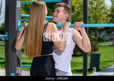 Junge attraktive Frau zieht sich auf der horizontalen Bar mit einem Trainer auf einem Straßensportplatz zum Workout hoch. Stockfoto