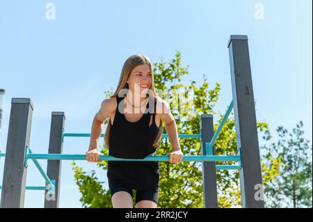 Ein junges Mädchen zieht sich auf einer horizontalen Bar auf einem Straßensportplatz zum Workout in einem Stadtpark hoch. Sonniger Sommertag. Stockfoto