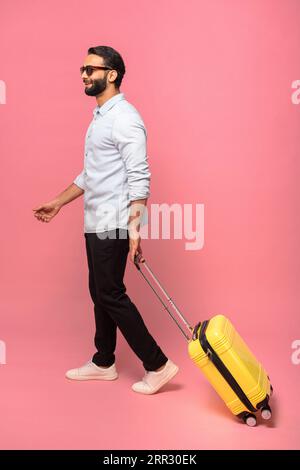 Vertikaler ganzer Blick auf den hübschen mittelalten indischen bärtigen Mann, der eine Sonnenbrille trägt und mit Gepäck läuft, den gelben Koffer auf rosa Hintergrund, geht in den Urlaub Stockfoto