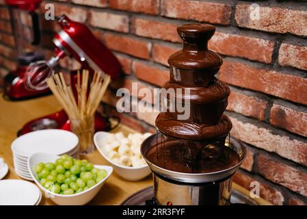 Schokoladenfonduenbrunnen in einem Topf neben einer roten Ziegelwand. Esstisch. Stockfoto