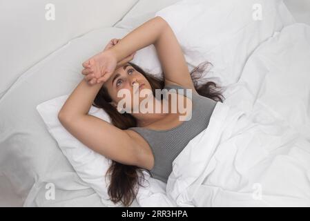 Junge Frau mit langen Haaren liegt im Bett und schaut auf die Schlafzimmerdecke, die unter Schlaflosigkeit leidet Stockfoto