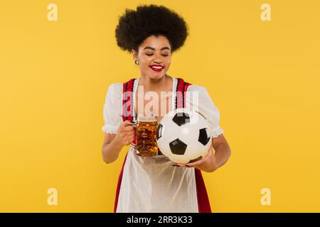 Fröhliche afroamerikanische oktoberfest-Kellnerin im Dirndl, die Bierbecher mit Fußball auf Gelb klirrrte Stockfoto