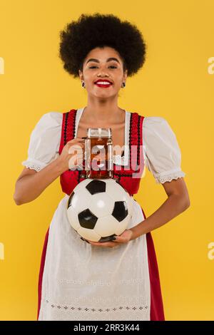 Fröhliche afroamerikanische bayerische Kellnerin mit Bierbecher und Fußball lächelnd auf gelb Stockfoto