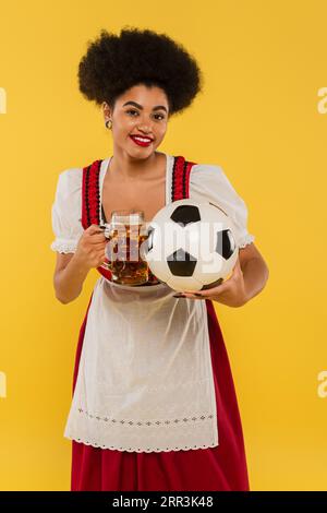 Fröhliche afroamerikanische oktoberfest-Kellnerin, die Bierbecher und Fußball auf Gelb klatscht Stockfoto