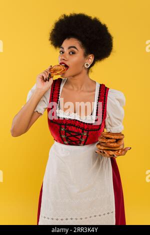 Charmante afroamerikanische Kellnerin in oktoberfest-Kleidung, die köstliche Brezel auf Gelb aß Stockfoto