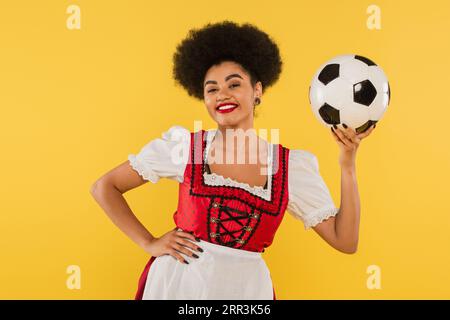 Charmante afroamerikanische bayerische Kellnerin mit Fußball und Hand auf Hüfte auf Gelb Stockfoto