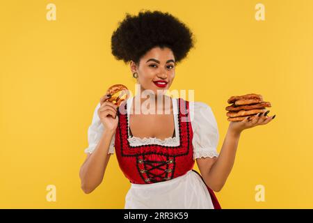 Fröhliche afroamerikanische Kellnerin im authentischen Kostüm, die Brezeln auf Gelb hält Stockfoto