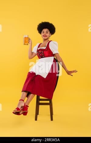 Fröhliche afroamerikanische Kellnerin im oktoberfest-Outfit, die auf einem Stuhl mit Bierbecher auf gelb sitzt Stockfoto