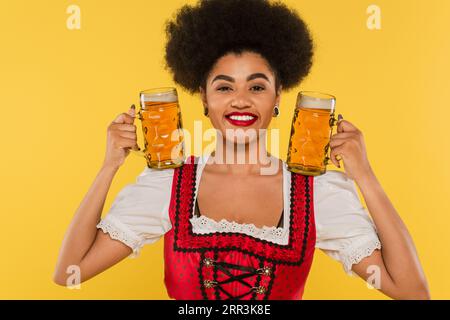 Lächelnde afroamerikanische oktoberfest-Kellnerin im Dirndlkleid mit Tassen mit Craft-Bier auf Gelb Stockfoto