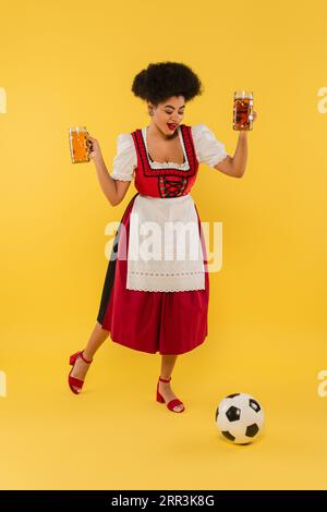 Fröhliche afroamerikanische oktoberfest-Kellnerin, die Bierbecher hält und Fußball auf Gelb spielt Stockfoto