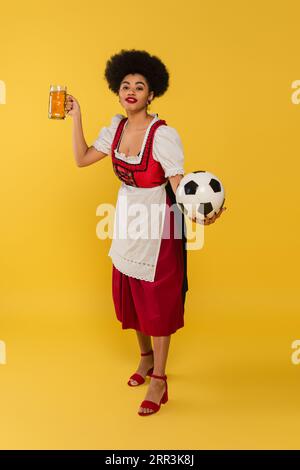 Freute afroamerikanische Kellnerin in bayerischer Tracht mit Bierbecher und Fußball auf gelb Stockfoto