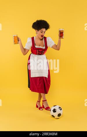 Fröhliche afroamerikanische bayerische Kellnerin im Dirndl, die Bierbecher in der Nähe des Fußballs auf gelb hält Stockfoto