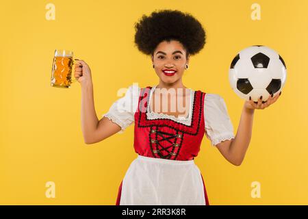 Fröhliche afroamerikanische bayerische Kellnerin im Dirndl mit Bierbecher und Fußball auf gelb Stockfoto