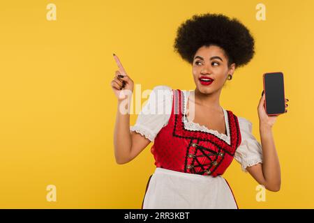 afroamerikanische bayerische Kellnerin mit Smartphone, das mit dem Finger zeigt und die Richtung auf gelb zeigt Stockfoto