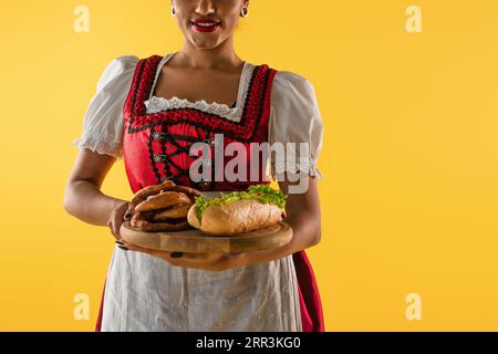 afroamerikanisch-bayerische Kellnerin mit Brezeln und Hot Dog auf Holztablett auf gelb Stockfoto