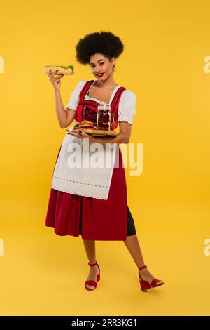 Fröhliche afroamerikanische oktoberfest-Kellnerin mit Bier und köstlichen Leckereien auf Holztablett auf gelb Stockfoto