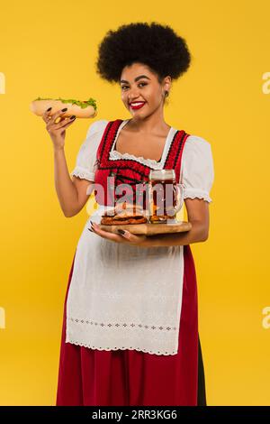 Fröhliche afroamerikanische bayerische Kellnerin mit Bier, Brezeln und Hot Dog auf Holztablett auf gelb Stockfoto