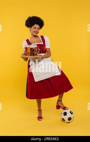 Fröhliche afroamerikanische oktoberfest-Kellnerin mit Bierbechern auf Holztablett in der Nähe von Fußball auf Gelb Stockfoto