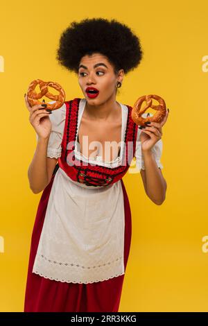 Staunte afroamerikanische Kellnerin im oktoberfest-Outfit mit köstlichen Brezeln auf Gelb Stockfoto