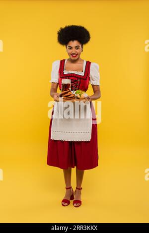 afroamerikanische oktoberfest-Kellnerin, die ein Holztablett mit Bier und köstlichen Leckereien auf gelb hält Stockfoto