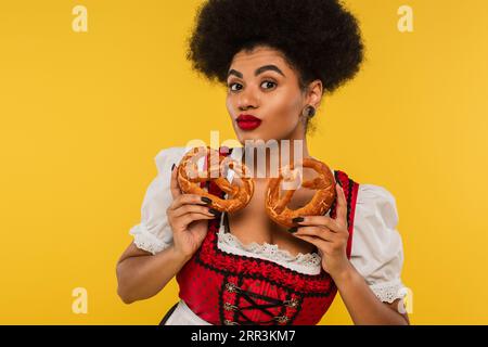 Charmante afroamerikanische oktoberfest-Kellnerin mit köstlichen Brezeln, die die Kamera auf Gelb betrachten Stockfoto