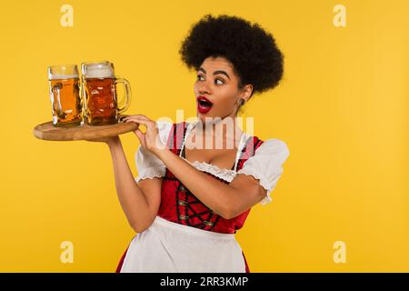 Staunte afroamerikanische oktoberfest-Kellnerin, die ein Holztablett mit Bierbechern auf gelbem Banner hielt Stockfoto