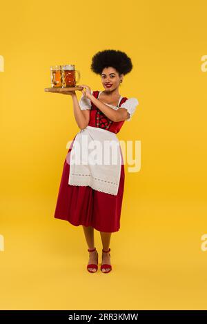 Lächelnde afroamerikanische oktoberfest-Kellnerin, die ein Holztablett mit Bierbechern auf gelbem Banner hält Stockfoto