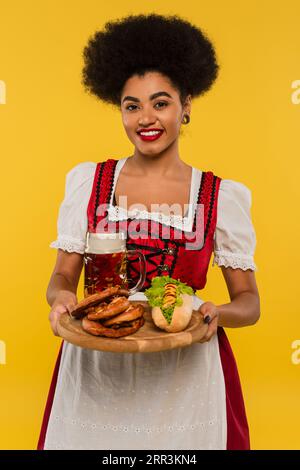 Fröhliche afroamerikanische oktoberfest-Kellnerin mit Holztablett mit Brezeln und Hot Dog auf gelb Stockfoto