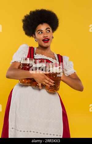 Fröhliche afroamerikanische bayerische Kellnerin im Dirndl, die Bierbecher hält und auf gelb schaut Stockfoto