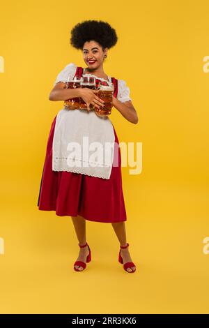 Fröhliche afroamerikanische oktoberfest-Kellnerin mit vollen Biermags, die die Kamera auf gelb anschauen Stockfoto