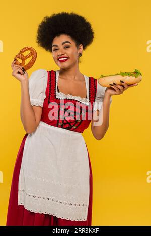 Hübsche afroamerikanische oktoberfest-Kellnerin mit Hotdog und Brezel lächelnd vor der Kamera auf gelb Stockfoto