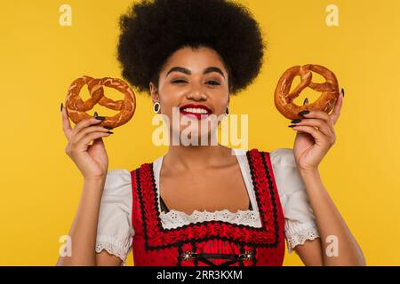 Fröhliche afroamerikanische oktoberfest-Kellnerin im bayerischen Dirndl mit leckeren Brezeln auf Gelb Stockfoto